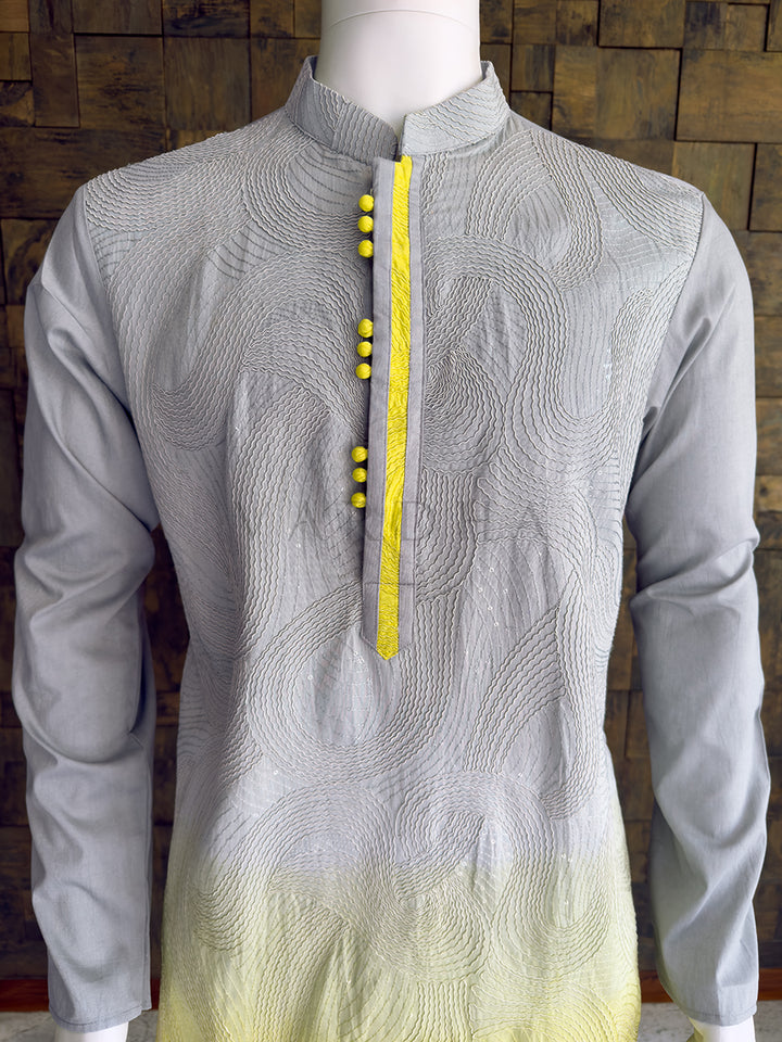 Gray kurta with yellow accents on a mannequin against a wooden paneled wall. Detailed chest view of the spherical yellow fabric buttons on the contrast placket, framed by swirling cord embroidery on the grey viscose mandarin collar kurta.