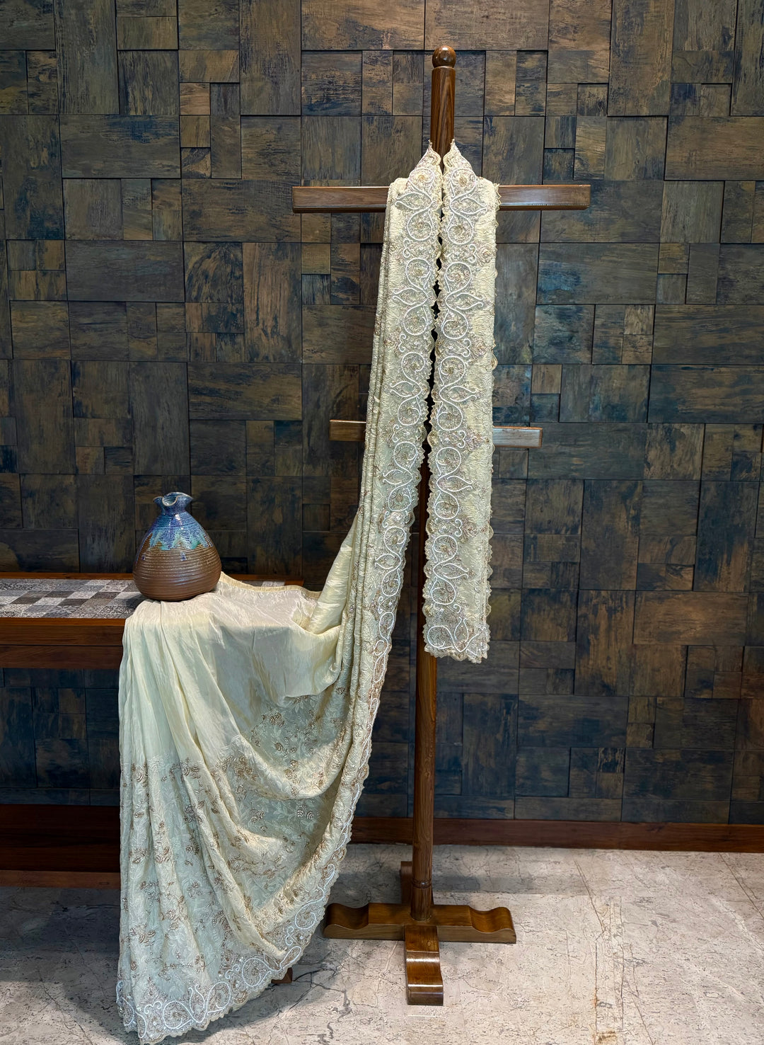 Ivory tissue silk saree draped on wooden stand showcasing intricate zardozi hand embroidery and scalloped border. Lace fabric draped over a wooden stand against a textured wall. 