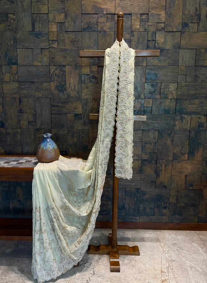 Ivory tissue silk saree draped on wooden stand showcasing intricate zardozi hand embroidery and scalloped border. Lace fabric draped over a wooden stand against a textured wall. 
