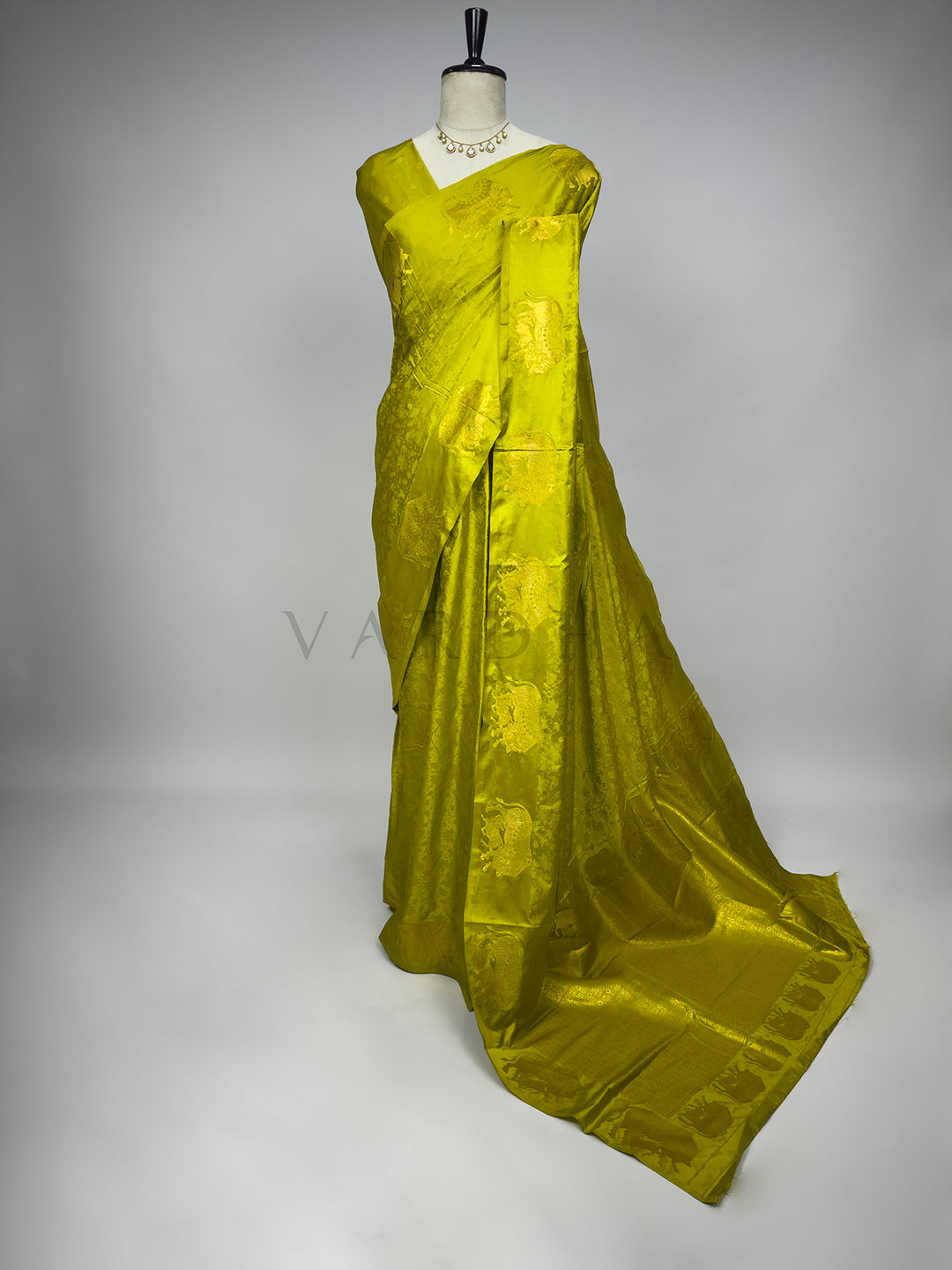Green saree on a mannequin against a gray background A full-length view of an elegant Leaf Green Satin Silk Saree beautifully draped on a mannequin, styled with a classic kundan necklace. The saree showcases a vibrant chartreuse-green body adorned with a subtle tone-on-tone floral jacquard texture, beautifully contrasted by large, majestic gold zari elephant motifs woven along the palla. The premium satin fabric cascades into a grand, fluid floor-length drape against a clean neutral background, perfect for