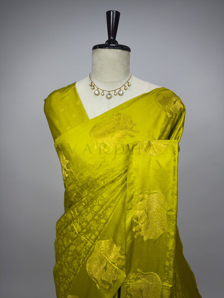 Green saree on a mannequin with a gray background A detailed half-torso perspective of the draped Leaf Green Satin Silk Saree on a mannequin, emphasizing the exquisite pattern layout over the chest and pleated shoulder drape. The glossy satin material highlights a delicate self-patterned floral vine design, while the shoulder section displays the intricately woven gold zari elephant motifs. Paired with a delicate stone statement necklace, this close view illustrates the regal charm of traditional Indian ha 