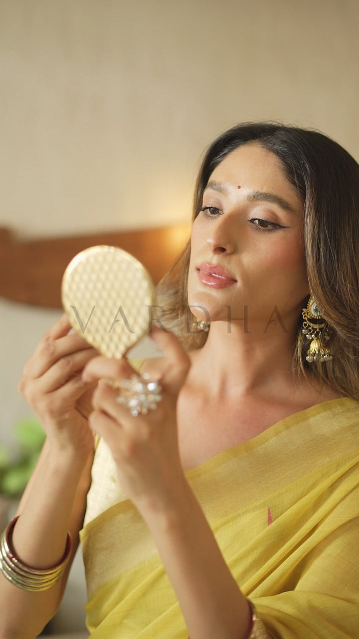 A woman in a yellow saree holds a small round compact mirror, looking at her reflection. She is wearing gold earrings and bangles, with a warm, softly lit indoor background. Beautiful model admiring herself in a hand mirror, showcasing the vibrant yellow hue and comfortable drape of the cotton jacquard festive saree.