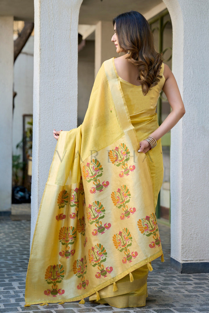 A woman wearing a yellow silk saree with orange and green floral motifs, standing in a corridor with white pillars. Back view of the lemon yellow cotton jacquard saree displaying the grand oversized woven Paithani peacock motifs alongside matching yellow fancy tassels on the designer pallu.