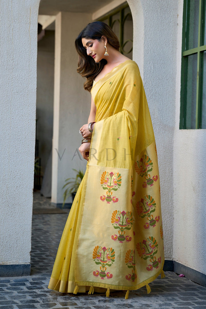 Woman wearing a yellow saree with floral patterns standing in front of a building. Stunning full-length draping of the vibrant yellow cotton jacquard handloom saree, showcasing the seamless blend of traditional Paithani weaves and perfect haldi color.