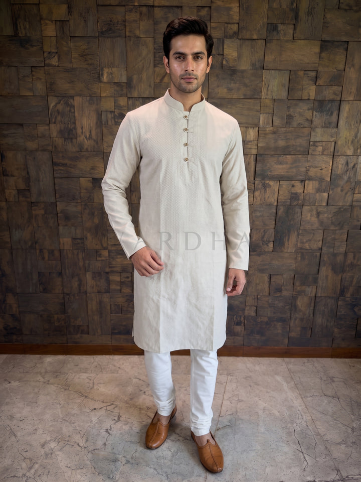 Man wearing a grey kurta with white pants against a wooden paneled wall. Light Grey men’s cotton kurta featuring a distinct woven micro-check texture that provides visual depth and a crisp, structured drape.