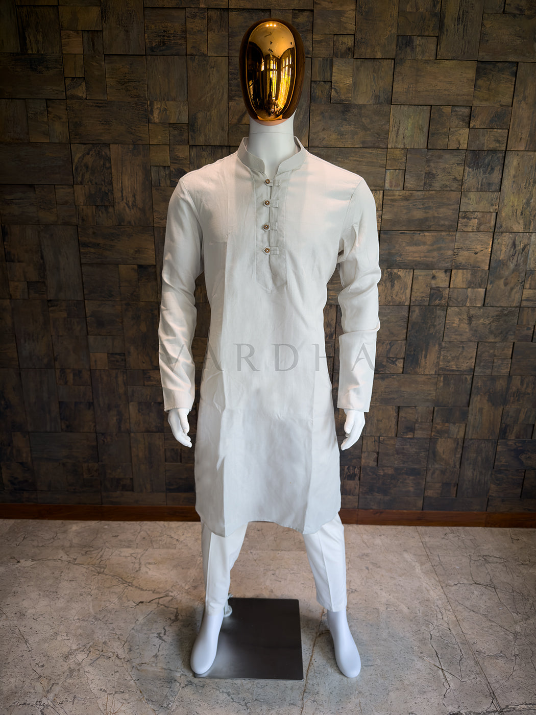 Mannequin wearing a grey traditional outfit with a gold mask against a wooden paneled wall. Modern minimalist ethnic wear: A knee-length ash grey cotton kurta with clean lines, a sharp Mandarin collar, and full sleeves.