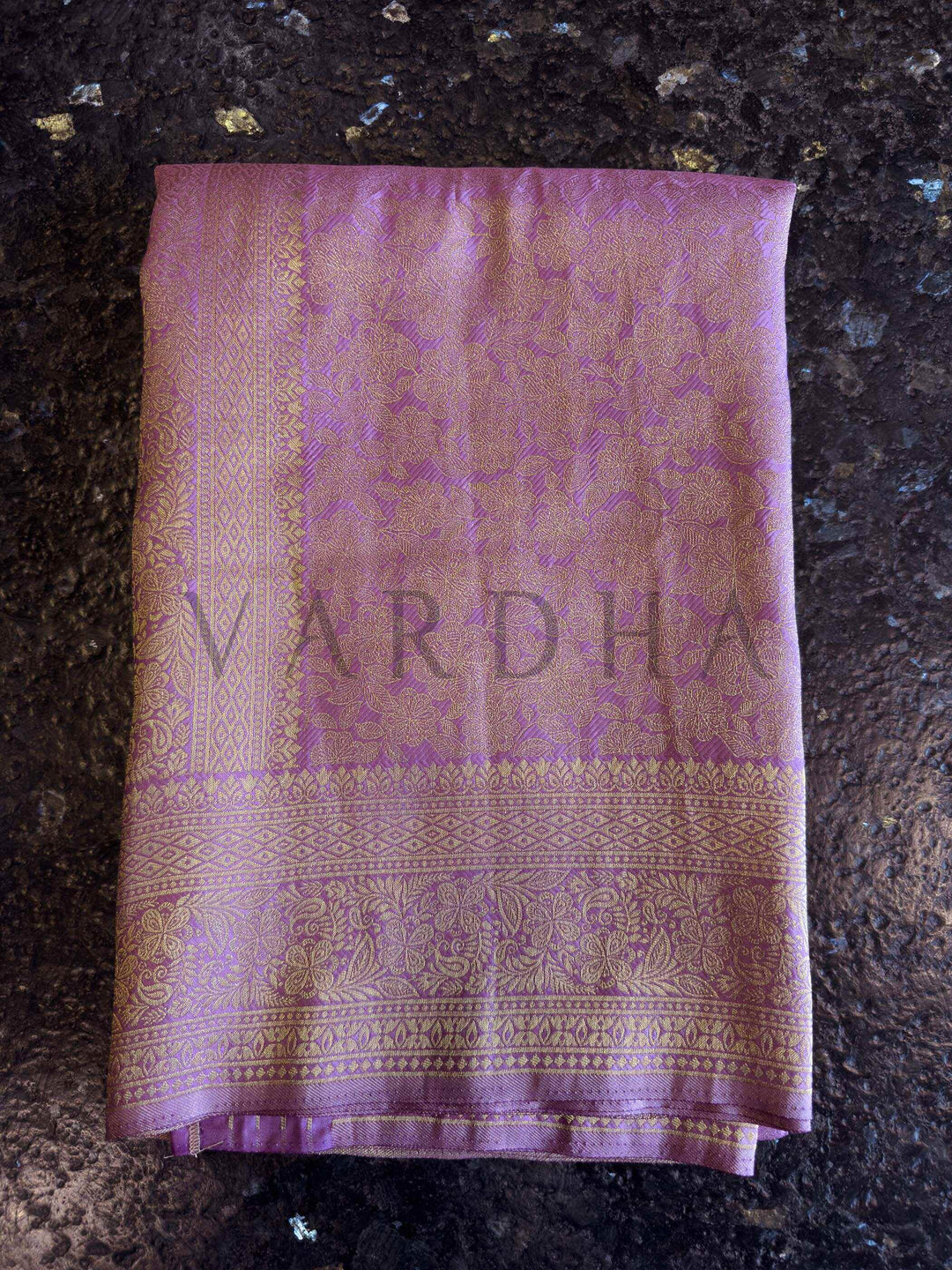 Macro texture shot of floral jaal weave on lavender saree showing intricate zari work and smooth fabric hand. Exclusive House of Vardha Lilac Kanjivaram Silk Saree displaying an elaborate Gold Zari Weave pallu and delicate floral motifs against a textured wall backdrop. We offer Ready to wear, Pre drape service, Saree Add on, Custom Stitched Blouse, Fall and Edging, Gift Packaging at Complimentary Price!