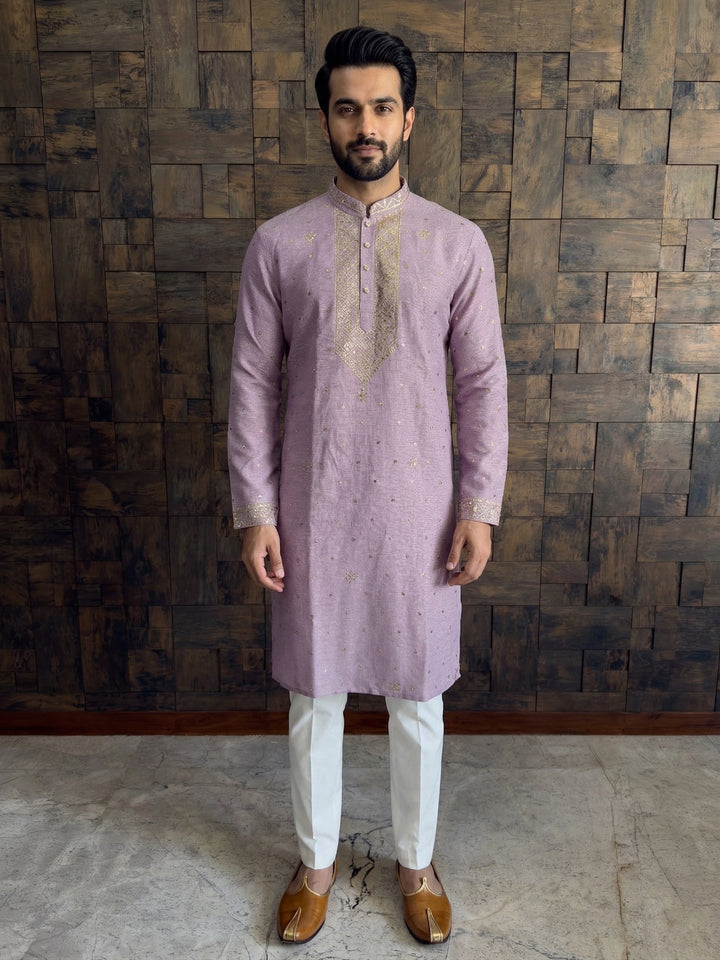 Man wearing a light purple kurta with white pants against a wooden paneled wall. Lilac khadi cotton kurta with gold floral mukaish work paired with white churidar and traditional mojari
