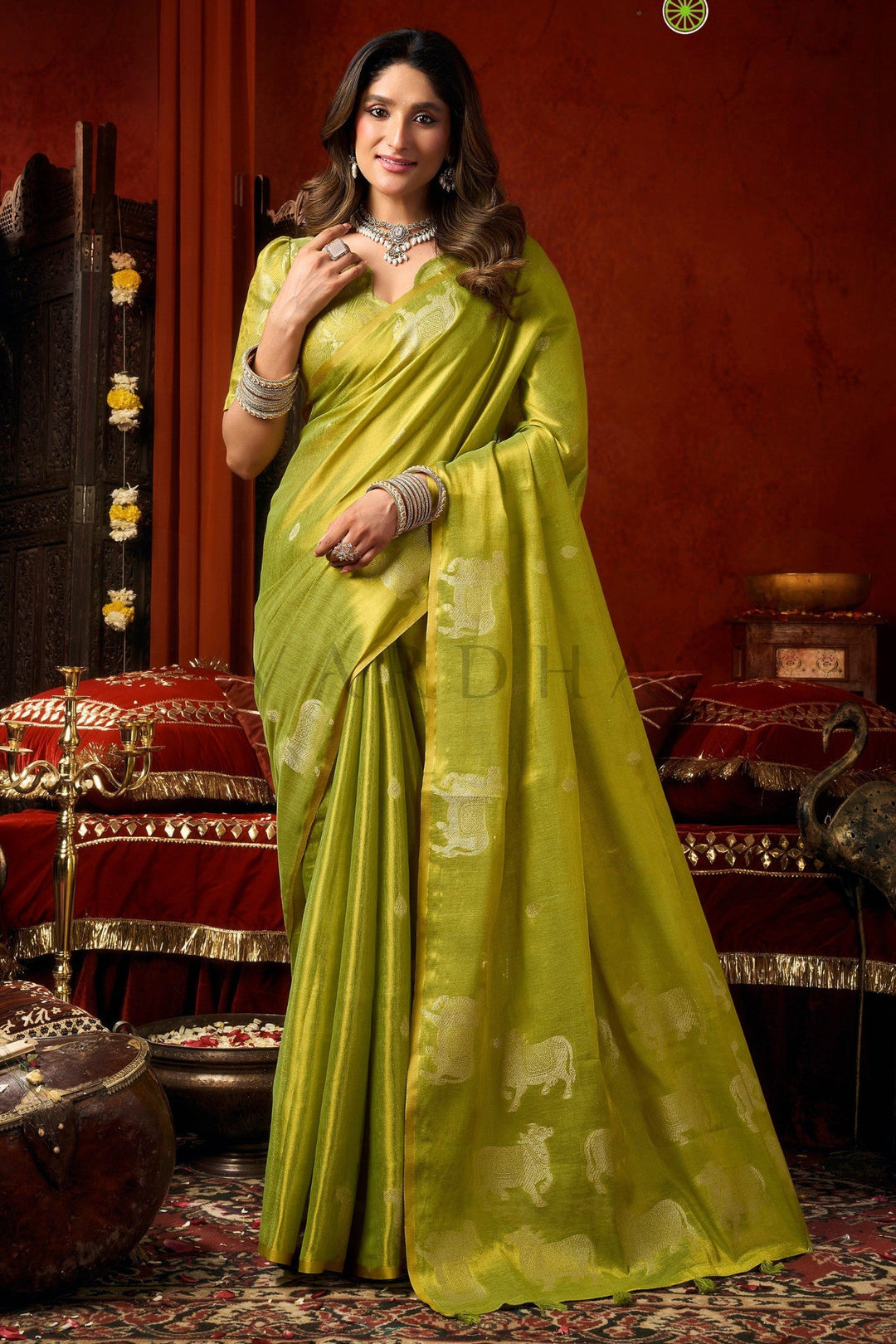 A woman wearing a bright yellow-green silk saree with light beige elephant motifs, posed in an ornate traditional room with red walls and decorative furniture. Full front view of an elegant lime green heavy tissue saree featuring traditional silver woven cow buttas and a gold zari border.