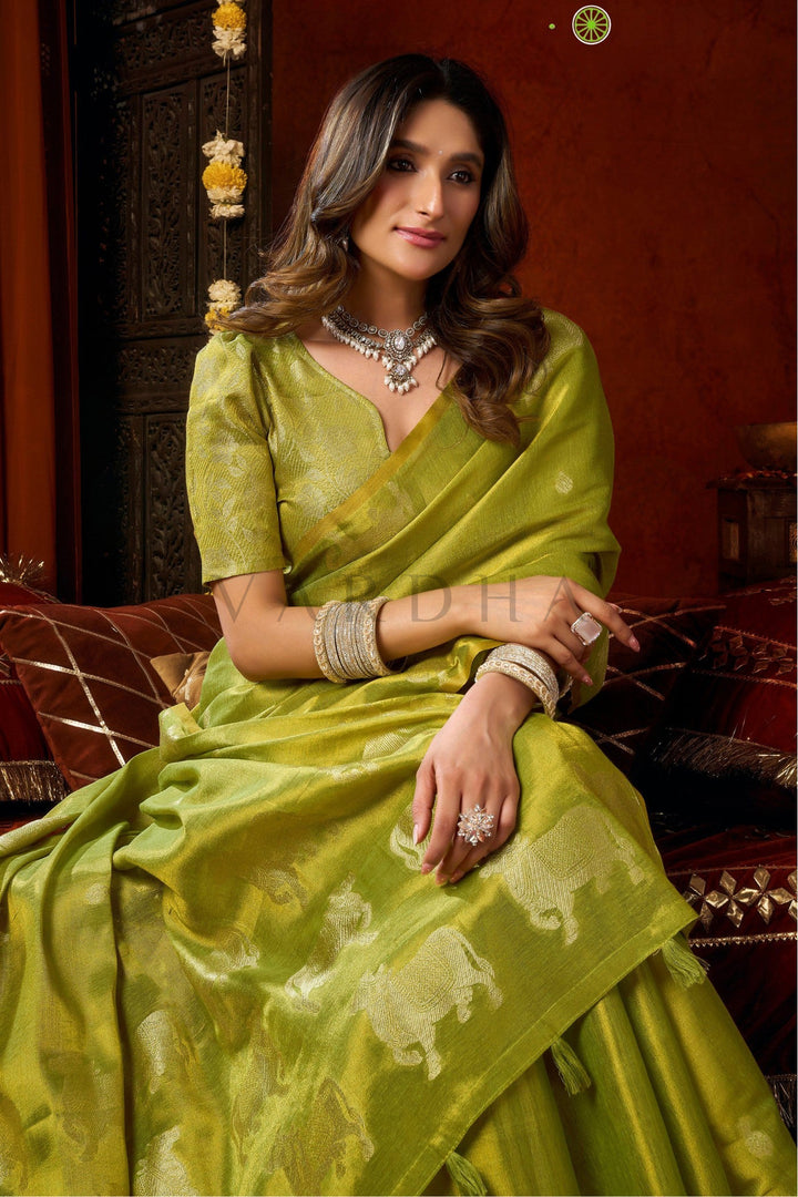 Woman in a green saree sitting on a decorative chair with a red wall background Beautiful woman sitting gracefully in a vibrant green tissue silk saree, showcasing the luxurious drape and matching woven blouse.