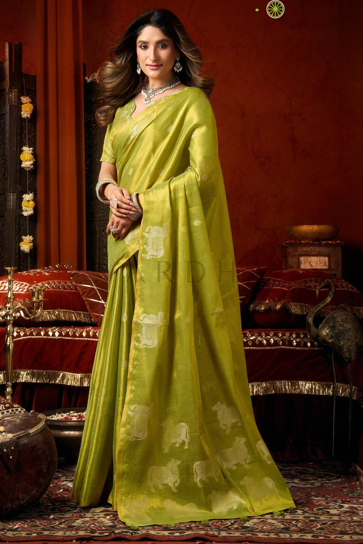 Woman wearing a green saree with a traditional setting Side standing view of the green festive wear heavy tissue saree highlighting the continuous Pichwai-style cow motifs and flawless fall.