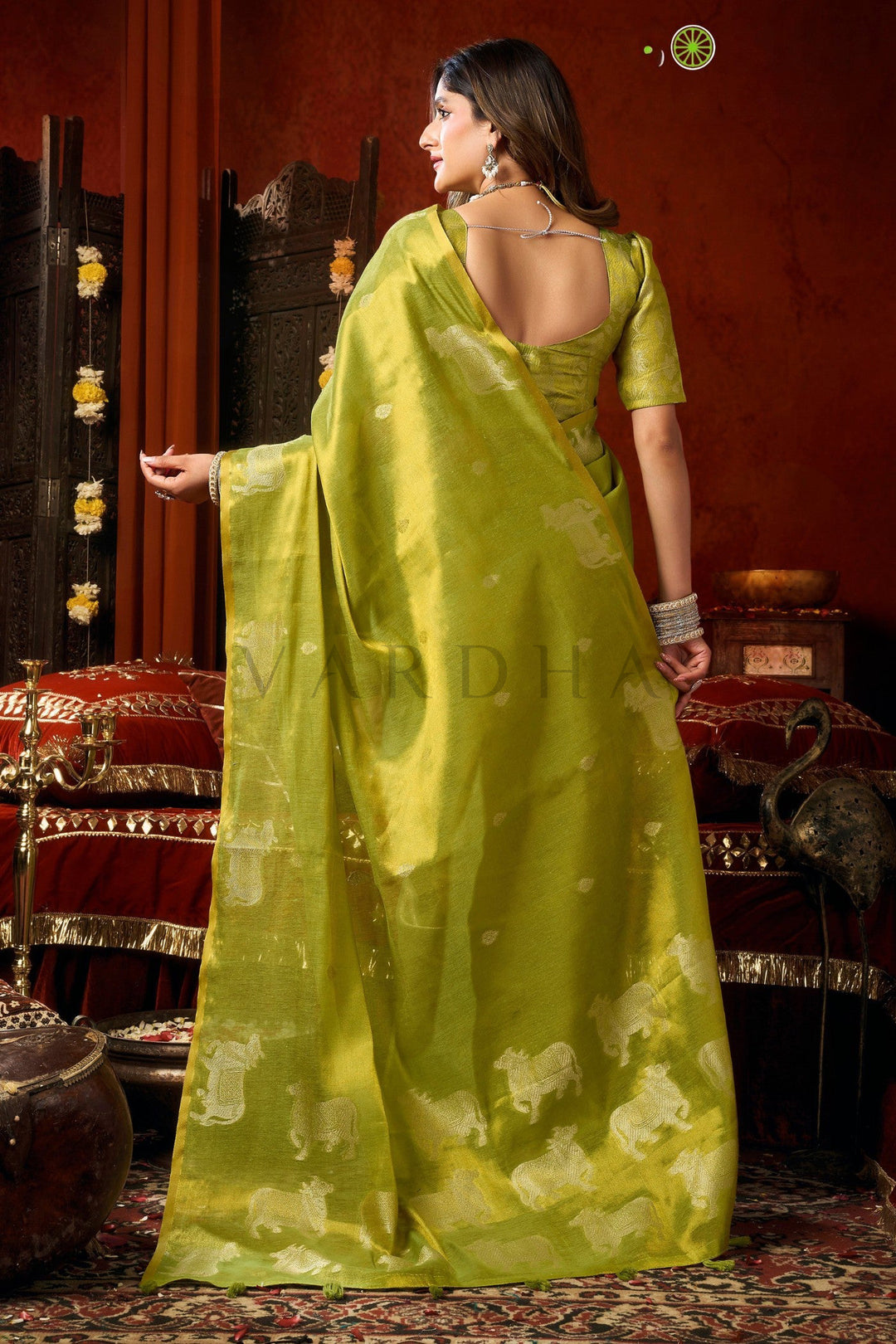 Woman in a green saree with a traditional setting Back view of the radiant green tissue saree, emphasizing the rich metallic sheen, woven silver cow designs, and elegant fancy tassels.