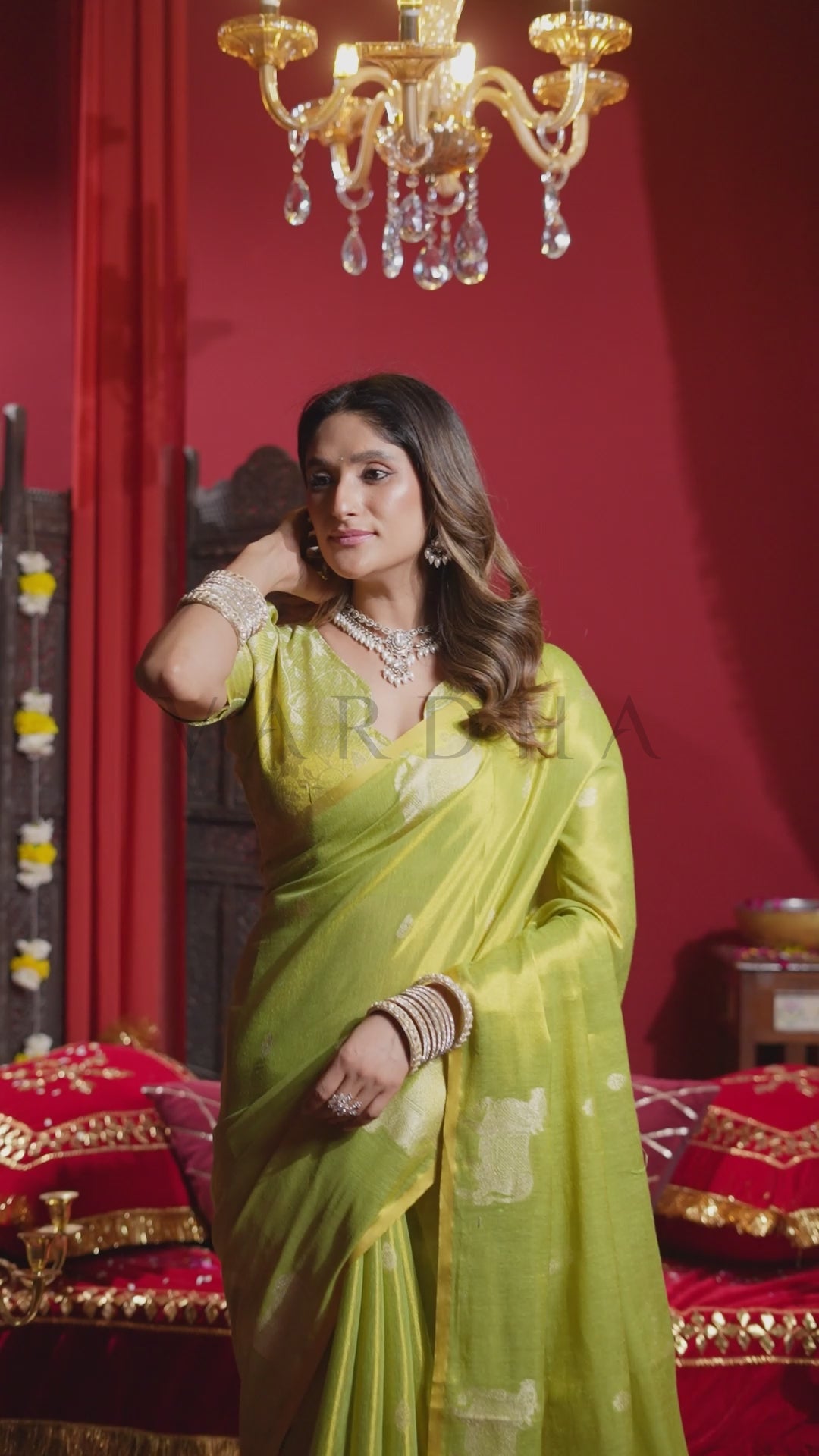 A woman wearing a light green saree with gold floral motifs, standing in a richly decorated room with red walls, a gold chandelier, and red upholstered furniture. Glamorous evening shot of a woman wearing the vibrant green heavy tissue saree, highlighting its regal metallic glow under chandelier lighting.