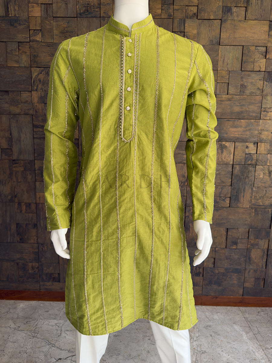 Green kurta on a mannequin against a wooden paneled wall. Close-up of the bright olive green men's festive kurta showcasing the textured silk-like drape, vertical zari stripes, and an intricately bordered button placket.