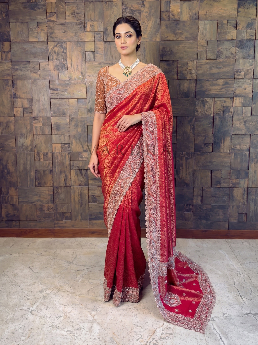 Woman wearing a red saree with a patterned border against a textured wall.