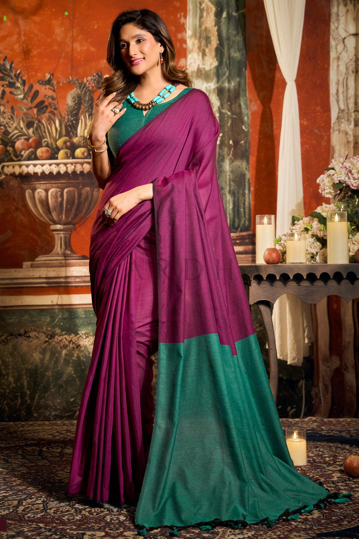 Woman wearing a purple and green saree in an indoor setting with decorative elements. Full front view of a woman wearing a vibrant magenta mal cotton saree featuring a stunning teal green contrast pallu and fancy tassels.
