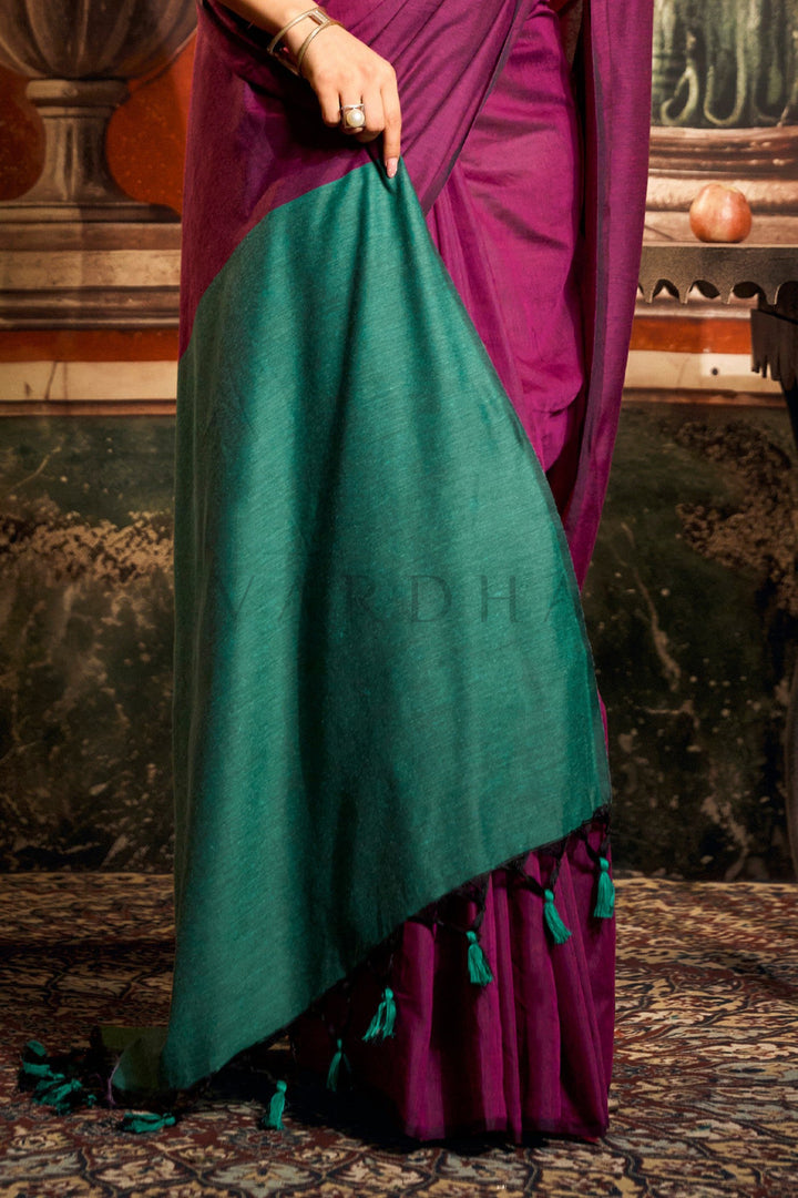 A close-up of a person wearing a draped sari in deep emerald green and rich magenta-purple, with matching tassel details, set against an ornate indoor background. Detailed fabric close-up of the magenta mal cotton saree highlighting the beautifully striking teal green contrast pallu edge with playful tassels.