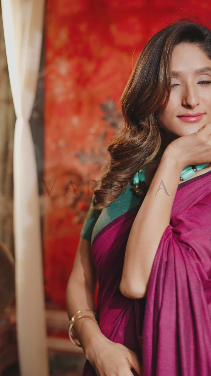 A woman wearing a magenta and teal sari, posing with her eyes closed against a warm red and orange patterned backdrop. Beautiful woman posing gracefully in an elegant jewel-toned magenta and teal dual-tone mal cotton saree, highlighting its minimalist and effortlessly chic aesthetic.