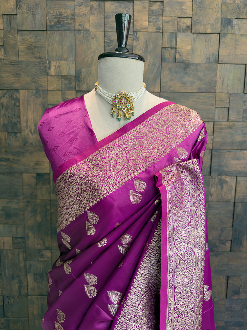 Purple saree with intricate patterns on a mannequin against a textured wall background Close-up of the magenta satin silk saree draped over the shoulder, featuring an intricate zari woven neckline and border.