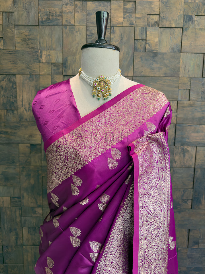 Purple saree with intricate patterns on a mannequin against a textured wall background Close-up of the magenta satin silk saree draped over the shoulder, featuring an intricate zari woven neckline and border.