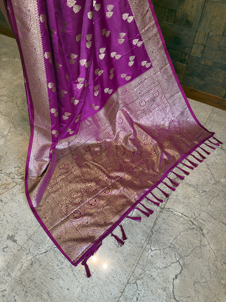 Purple and pink saree with intricate patterns on a tiled floor. Close-up highlighting the golden leaf bootis and dense floral woven border on the luxurious magenta satin fabric. Lower pleats and fluid, glossy finish of the magenta Banarasi-style satin silk ethnic saree.
