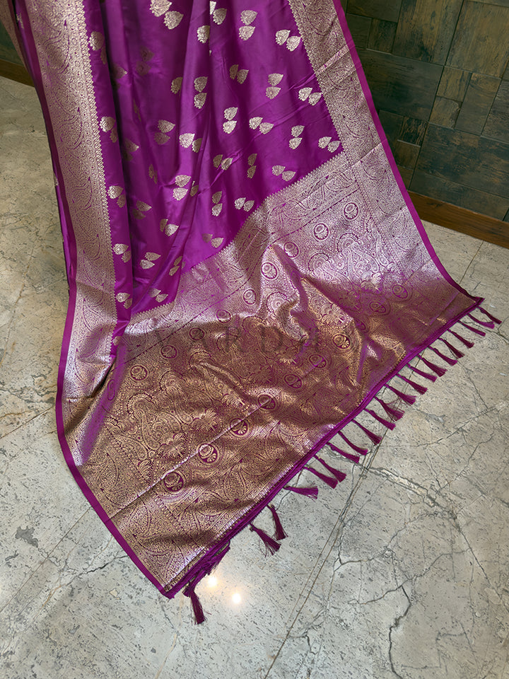 Purple and pink saree with intricate patterns on a tiled floor. Close-up highlighting the golden leaf bootis and dense floral woven border on the luxurious magenta satin fabric. Lower pleats and fluid, glossy finish of the magenta Banarasi-style satin silk ethnic saree.