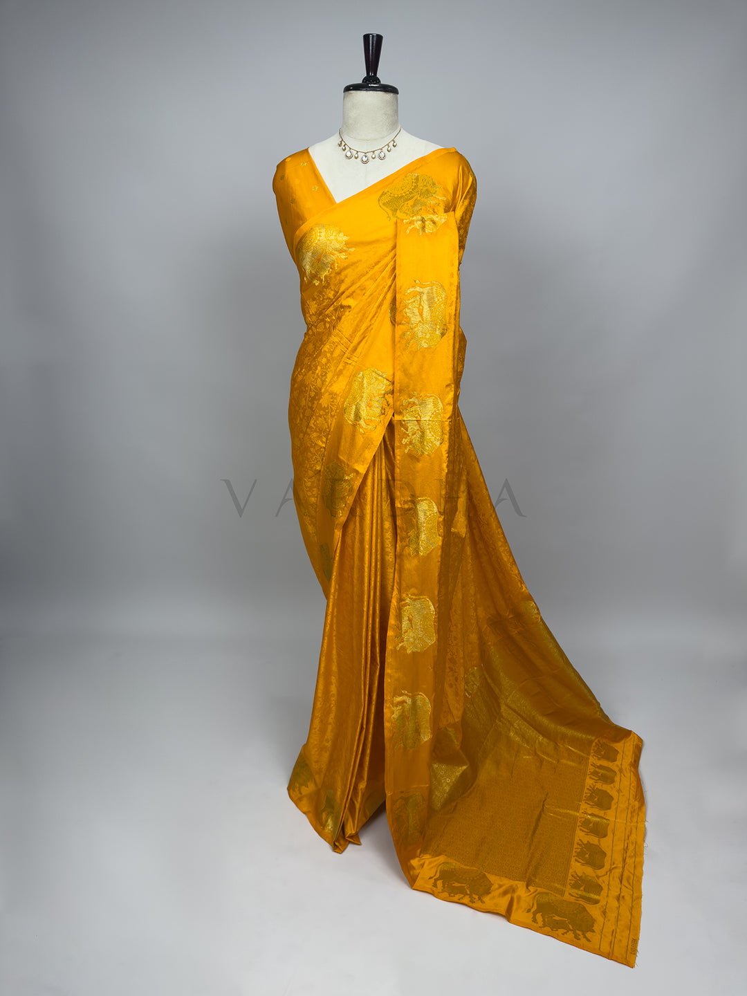 Yellow saree with a white blouse on a mannequin against a gray background A full-length drape of an Exquisite Marigold Orange Satin Silk Saree on a mannequin styled with a delicate kundan necklace. The saree features a radiant golden-orange hue with large, prominent gold zari elephant motifs woven beautifully along the palla and border. The smooth satin silk creates rich, fluid folds that extend into a majestic floor-length drape, showcasing a luxurious and royal ensemble perfect for traditional weddings an