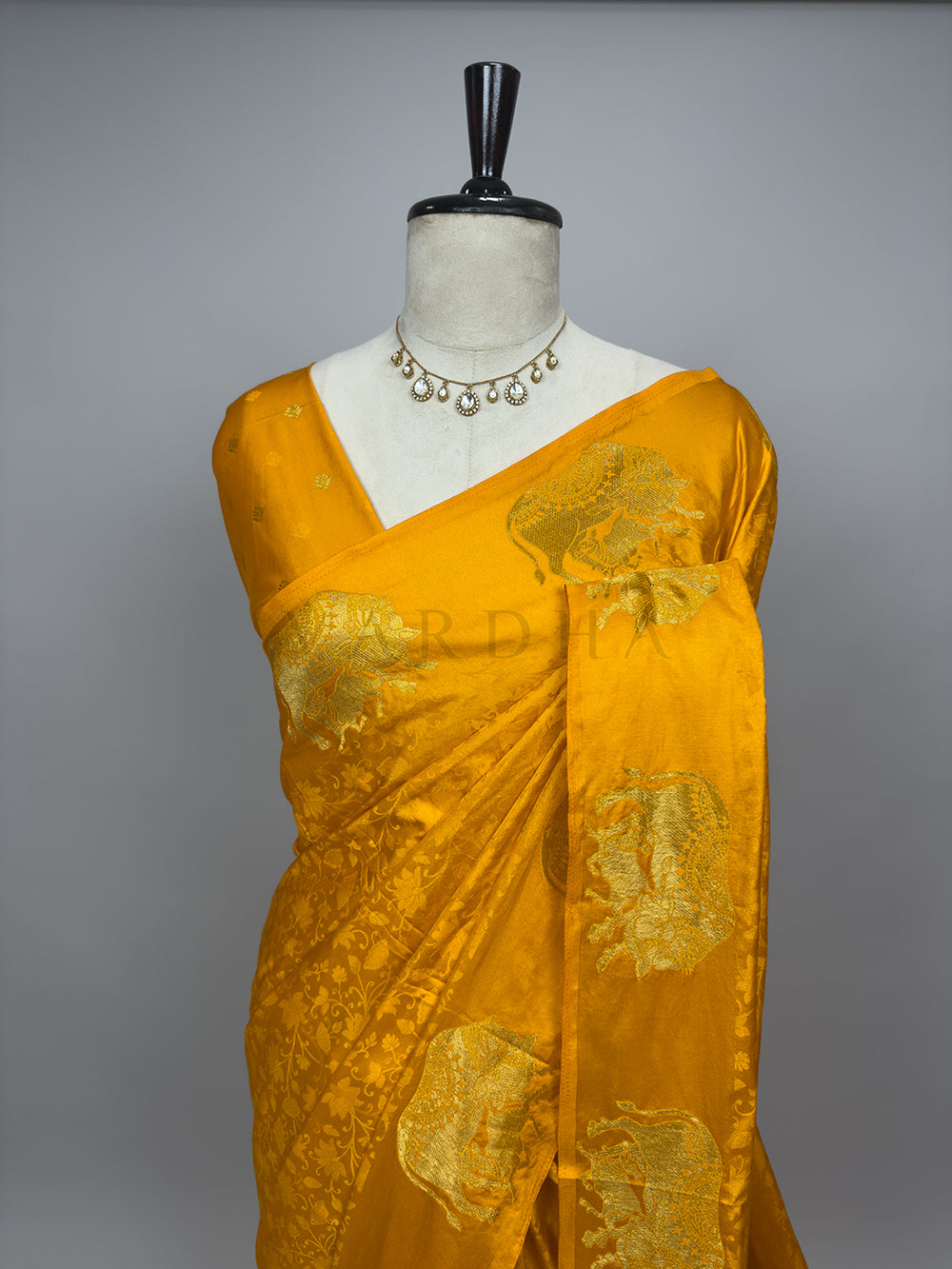 Yellow saree with gold patterns on a mannequin against a gray background A closer half-torso view of the draped Marigold Orange Satin Silk Saree on a mannequin, highlighting the intricate gold zari motifs across the chest and pleated shoulder drape. The ornate elephant designs stand out prominently against the brilliant orange background, demonstrating the premium quality of the fabric. Combined with a classic statement necklace, this angle displays the elegant silhouette and bridal styling potential of the