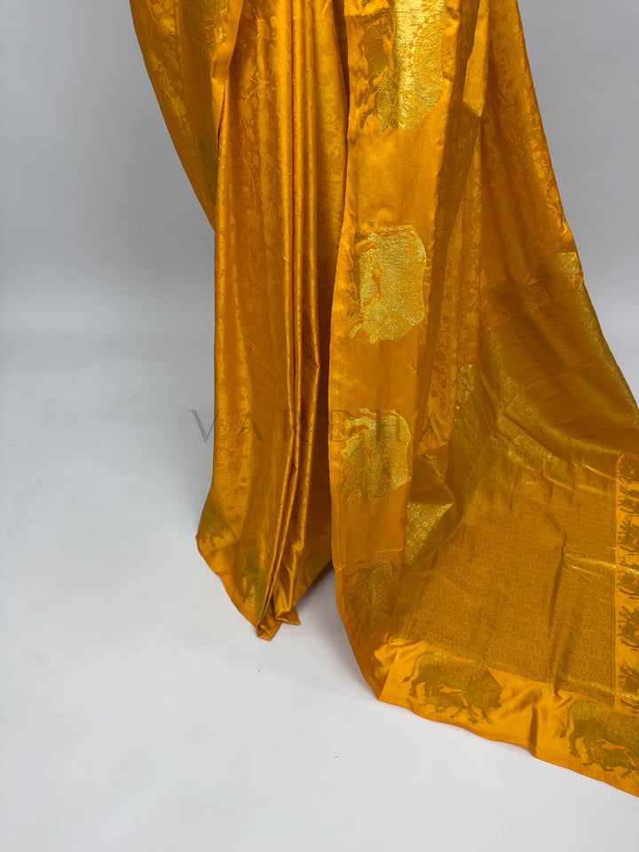 Gold fabric with intricate patterns on a white background A focused view of the magnificent spread-out pallu section of the Marigold Orange Satin Silk Saree. The shot captures the heavy, intricate gold zari weaving that blankets the palla in a dense geometric pattern, neatly bordered by an exquisite parade of marching woven elephants. The shimmering metallic threads against the rich orange background illustrate the high-end craftsmanship and heritage style of the Banarasi weave.