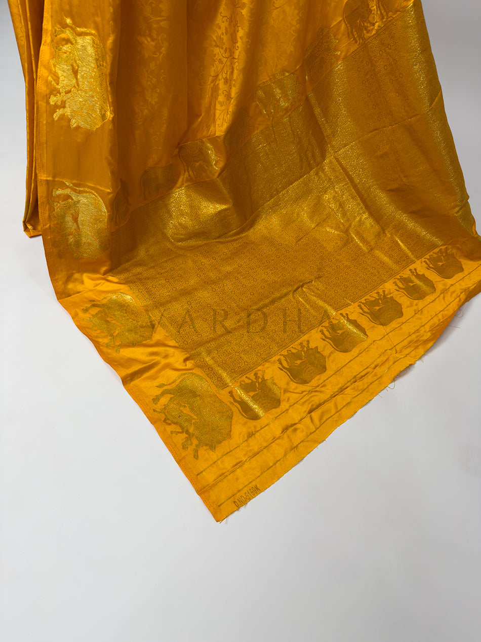 Gold fabric with embossed text and patterns on a white background An extreme close-up detail of the Marigold Orange Satin Silk fabric, showcasing the fine weave and luxurious texture in sharp focus. The image reveals a subtle self-patterned floral jacquard design woven directly into the vibrant orange satin base, overlapping with the shimmering metallic threads of the gold zari embroidery, emphasizing the premium quality and authentic artistry of this traditional handloom textile.