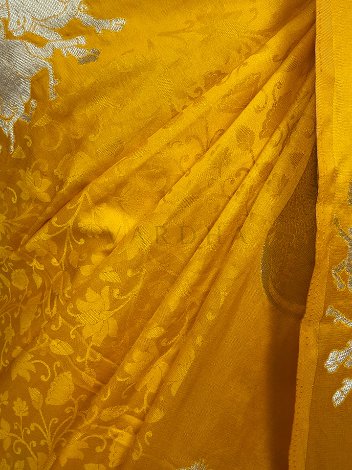 Close-up of yellow fabric with intricate patterns and embroidery. A targeted look at the lower section of the Marigold Orange Satin Silk Saree, focusing on the soft vertical folds and the graceful fall of the fabric. The smooth satin finish catches the light, accentuating the vibrant marigold color, while the vertical border exhibits a repeating line of meticulously woven gold zari elephant motifs that frame the traditional Indian attire beautifully.