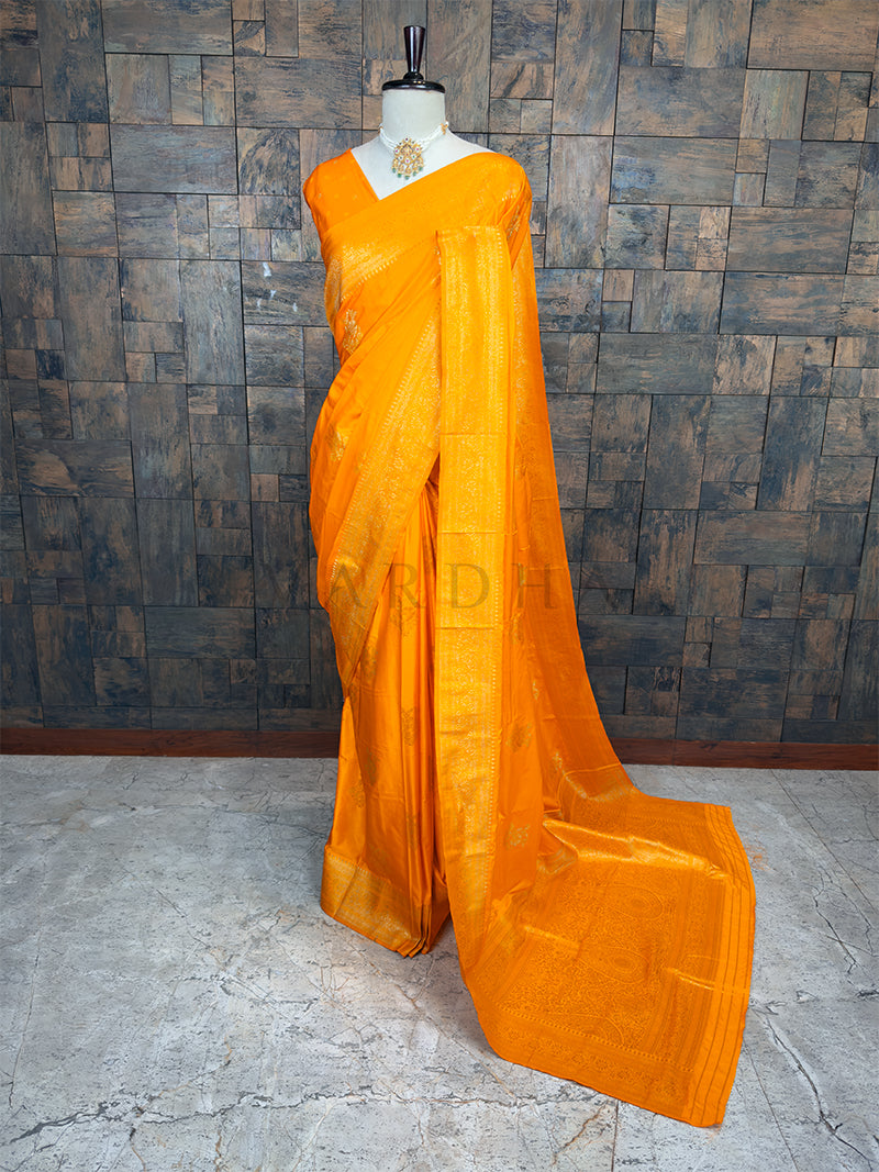Orange saree on a mannequin against a textured wall. House of Vardha Marigold Yellow Satin Saree featuring Antique Gold Zari Floral Motifs and a matching border, elegantly draped on a mannequin. We offer Ready to wear, Pre drape service, Saree Add on, Custom Stitched Blouse, Fall and Edging, Gift Packaging at Complimentary Price!