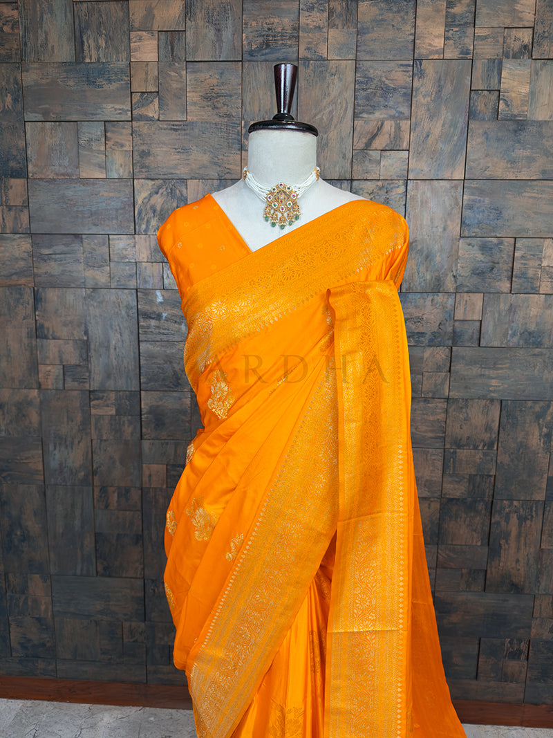 Orange saree with gold patterns on a mannequin against a wooden paneled wall. Lustrous Marigold Yellow Satin Saree by House of Vardha showcasing intricate Antique Gold Zari weaving and a vibrant, smooth texture. We offer Ready to wear, Pre drape service, Saree Add on, Custom Stitched Blouse, Fall and Edging, Gift Packaging at Complimentary Price!