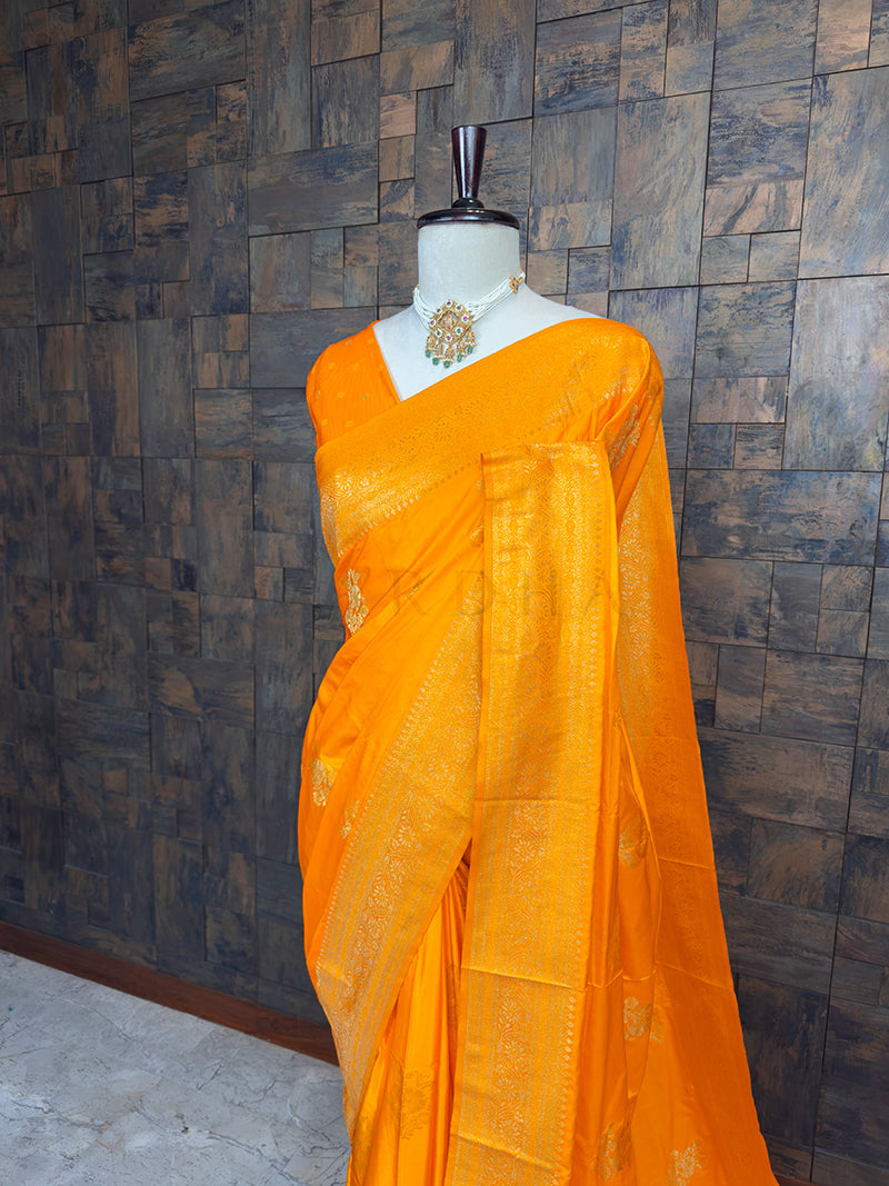 Orange saree on a mannequin against a wooden paneled wall. Close-up view of the House of Vardha Marigold Yellow Satin Saree highlighting the detailed Antique Gold Zari floral motifs and the beautifully woven traditional border. We offer Ready to wear, Pre drape service, Saree Add on, Custom Stitched Blouse, Fall and Edging, Gift Packaging at Complimentary Price!