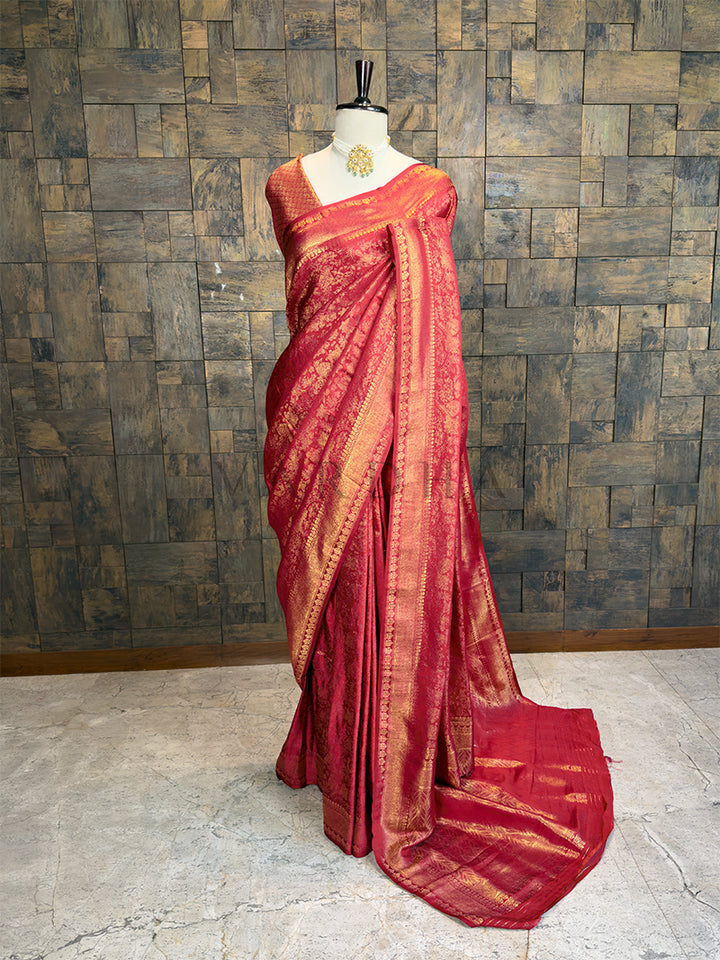 House of Vardha premium deep pinkish-red silk saree showcasing all-over gold zari brocade work draped on a mannequin. We offer Ready to wear, Pre drape service, Saree Add on, Custom Stitched Blouse, Fall and Edging, Gift Packaging at Complimentary Price!. Red saree with gold patterns on a mannequin against a textured wall.