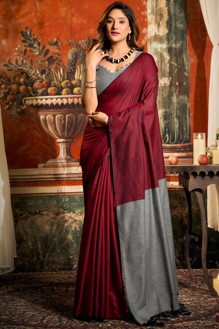 Woman wearing a maroon and gray saree in an indoor setting with decorative elements. Full front view of a woman wearing an elegant maroon mal cotton saree featuring a grey contrast pallu and fancy tassels.