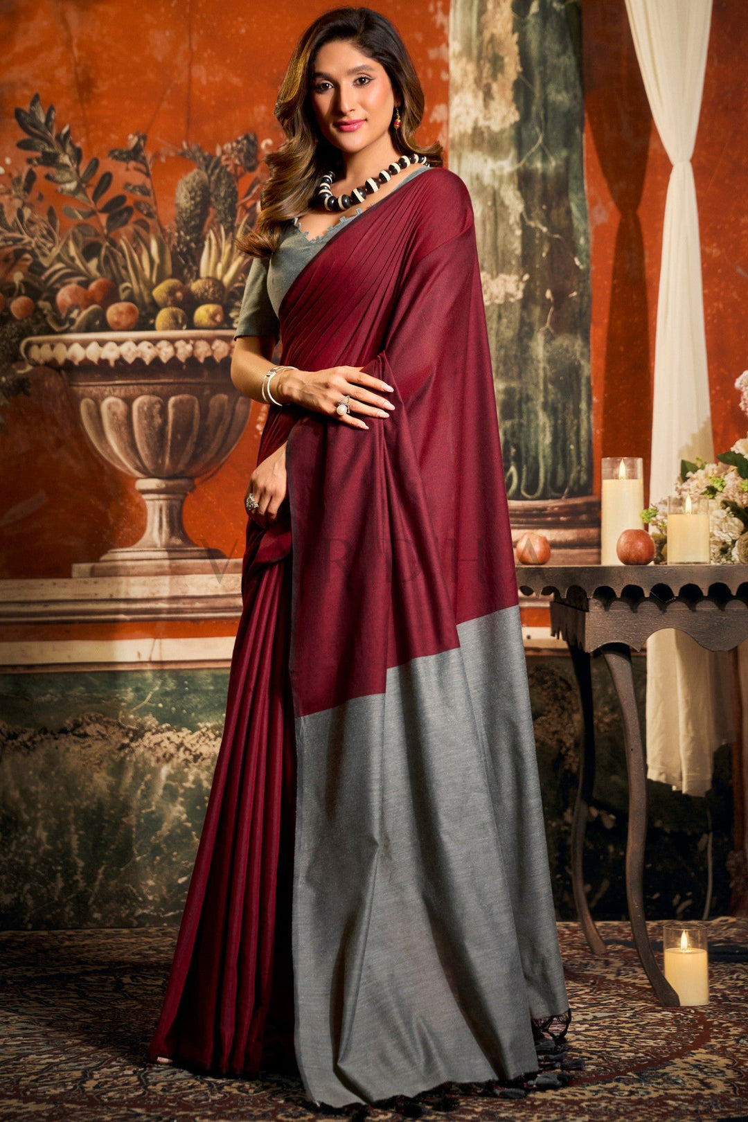 Woman wearing a maroon and gray saree in an elegant indoor setting. Stylish front drape of the lightweight dark red mal cotton saree, highlighting the striking grey contrast pallu for casual cultural outings.