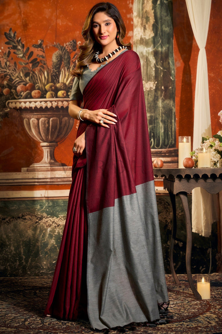 Woman wearing a maroon and gray saree in an elegant indoor setting. Stylish front drape of the lightweight dark red mal cotton saree, highlighting the striking grey contrast pallu for casual cultural outings.