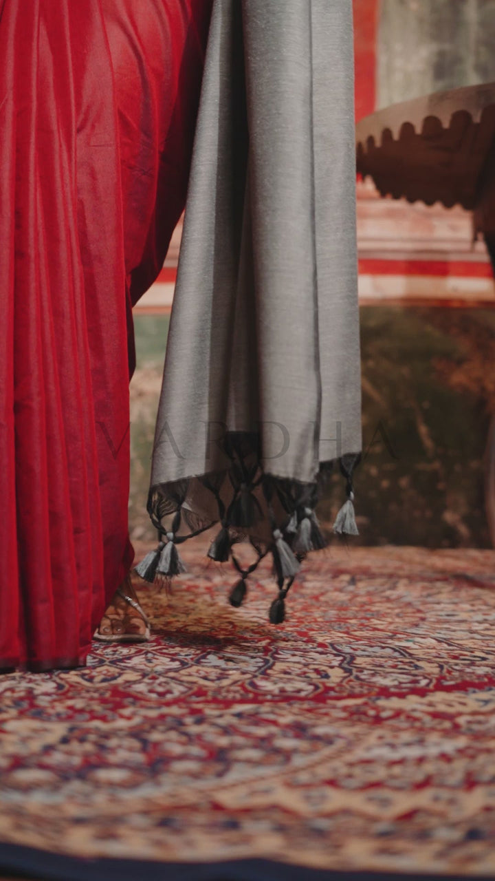A close-up of a gray shawl with black tassels draped over a red garment, resting on a patterned rug. Detailed fabric close-up of the maroon mal cotton saree highlighting the perfectly pleated texture and the solid grey contrast pallu edge.