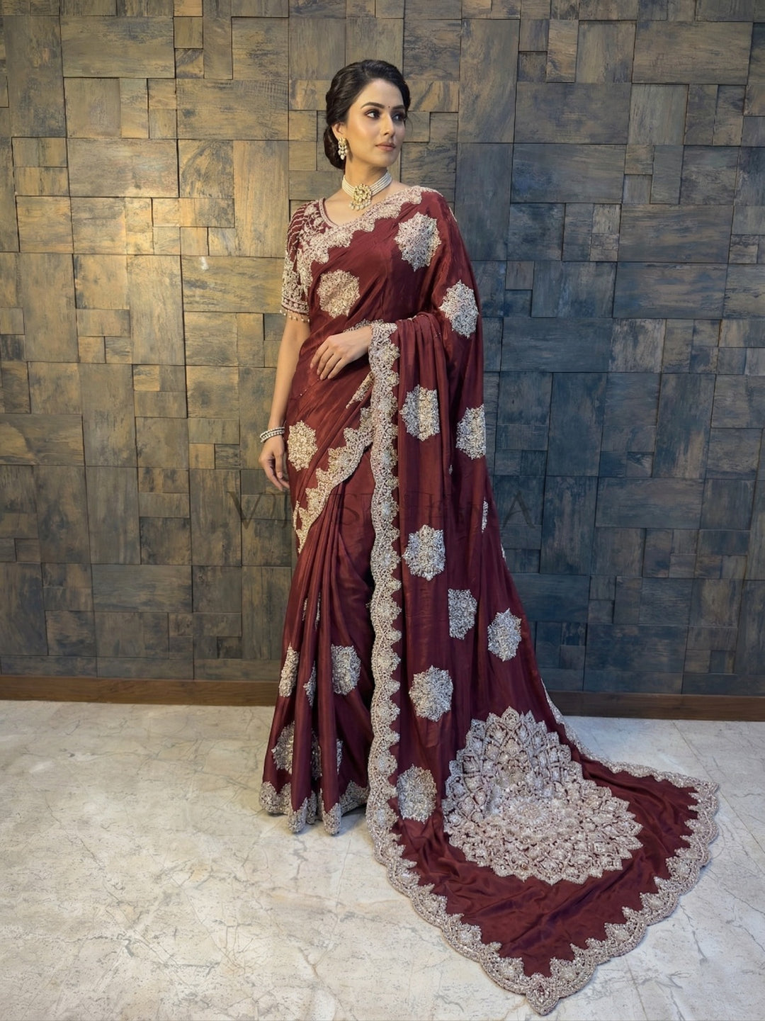 Woman in a maroon saree with white floral patterns against a textured wall. Maroon Satin Crepe Saree with Silver Cutdana & Beads Handwork