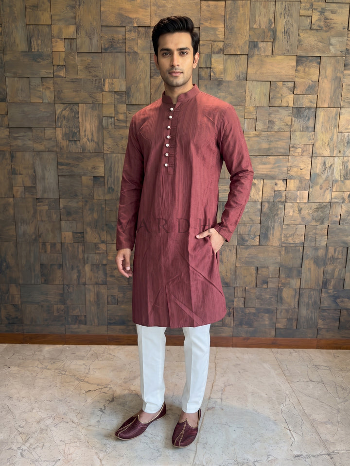 Man wearing a maroon kurta with white pants against a wooden paneled wall. Deep Maroon cotton kurta for men featuring a woven vertical micro-stripe dobby texture designed to add visual depth and elongate the silhouette.