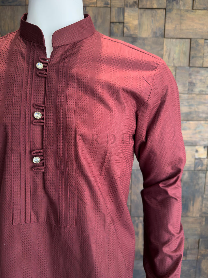 Maroon kurta on a mannequin against a brick wall background tailored with a clean Mandarin collar. The centerpiece of the design is the meticulously crafted chest placket, which is framed by sharp, vertical pleats (pintucks) that add a structured, formal "bib" effect. The placket is secured with traditional fabric button loops (tabs) and finished with premium, crystal-studded designer buttons that provide a luxurious, eye-catching sparkle against the dark maroon fabric.