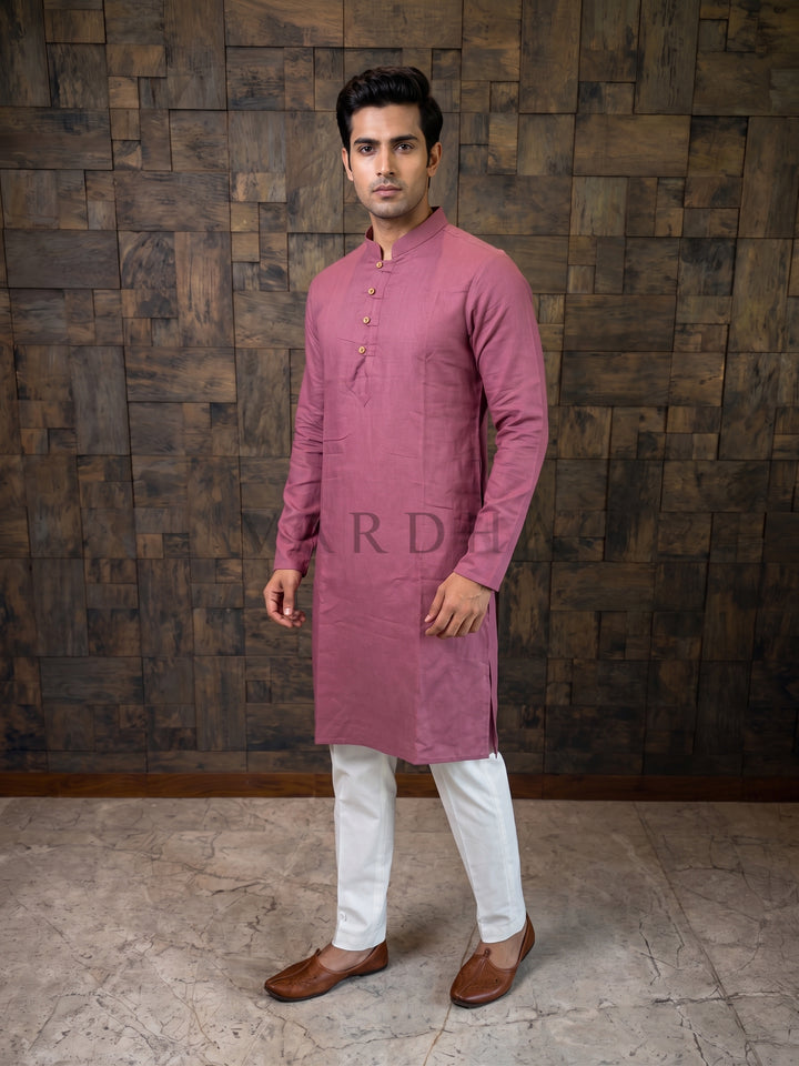 Man wearing a pink kurta with white pants against a wooden paneled wall. A knee-length Mauve Pink cotton kurta for men featuring a woven micro-check texture and an extended chest placket with traditional fabric button loops.