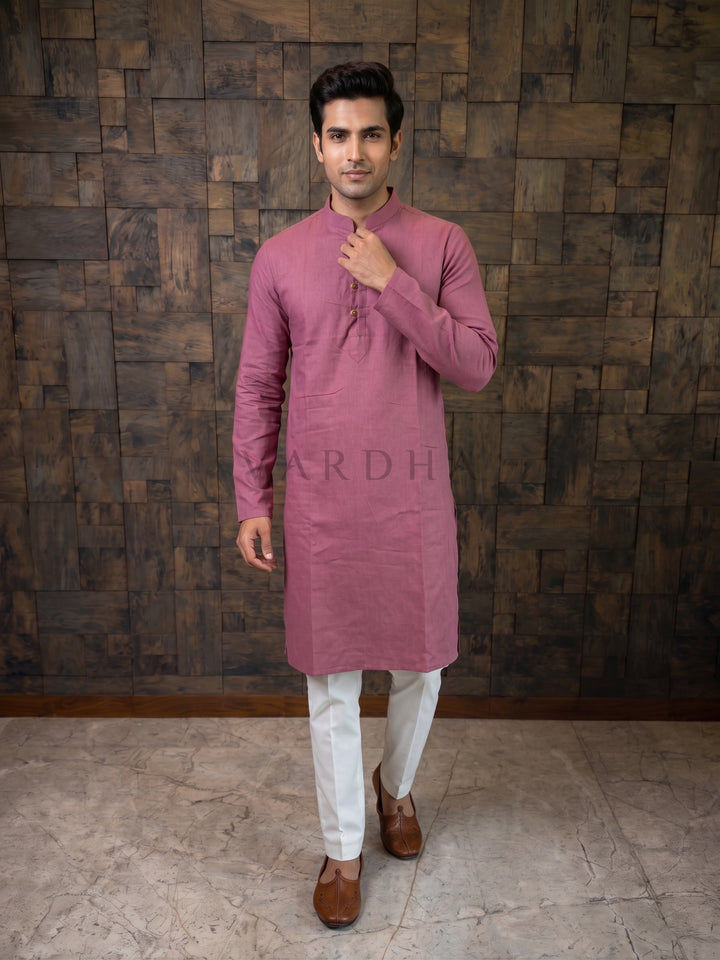 Man wearing a pink kurta with white pants against a wooden paneled wall. Detailed view of a designer dusty magenta kurta showing rustic, wooden-style buttons and artisanal fabric loops on a structured chest placket.