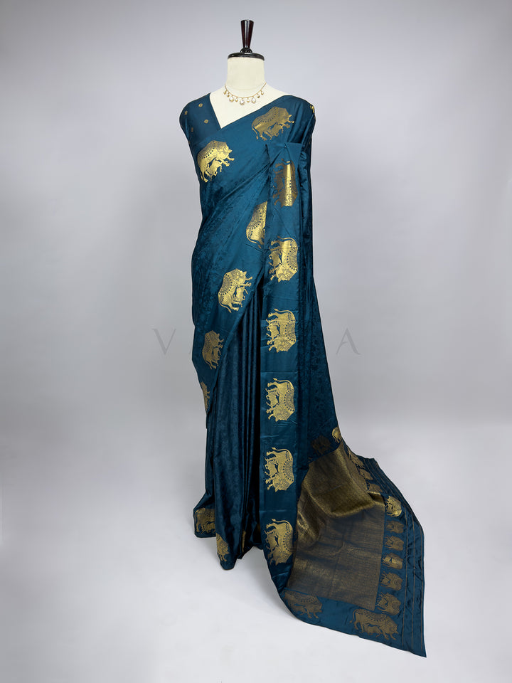 Blue saree with gold patterns on a mannequin against a white background A full-length presentation of a Midnight Blue Satin Silk Saree gracefully draped on a mannequin against a neutral gray background. The saree features a deep teal-blue body enhanced with a subtle self-jacquard floral pattern, contrasted beautifully by large, ornate gold zari elephant motifs along the outer drape. The heavy, richly woven pallu pools onto the floor, showcasing a majestic and royal ethnic look perfect for bridal wear and fe