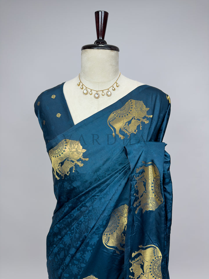 Blue saree with gold embroidery on a mannequin against a gray background A close-up torso view of the Midnight Blue Satin Silk Saree draped on a mannequin adorned with a delicate stone necklace. This angle highlights the fine details of the fabric, including the embossed tone-on-tone floral vines woven into the deep blue base and the prominent, shimmering gold zari elephant motifs on the chest and shoulder pleats, demonstrating premium craftsmanship.