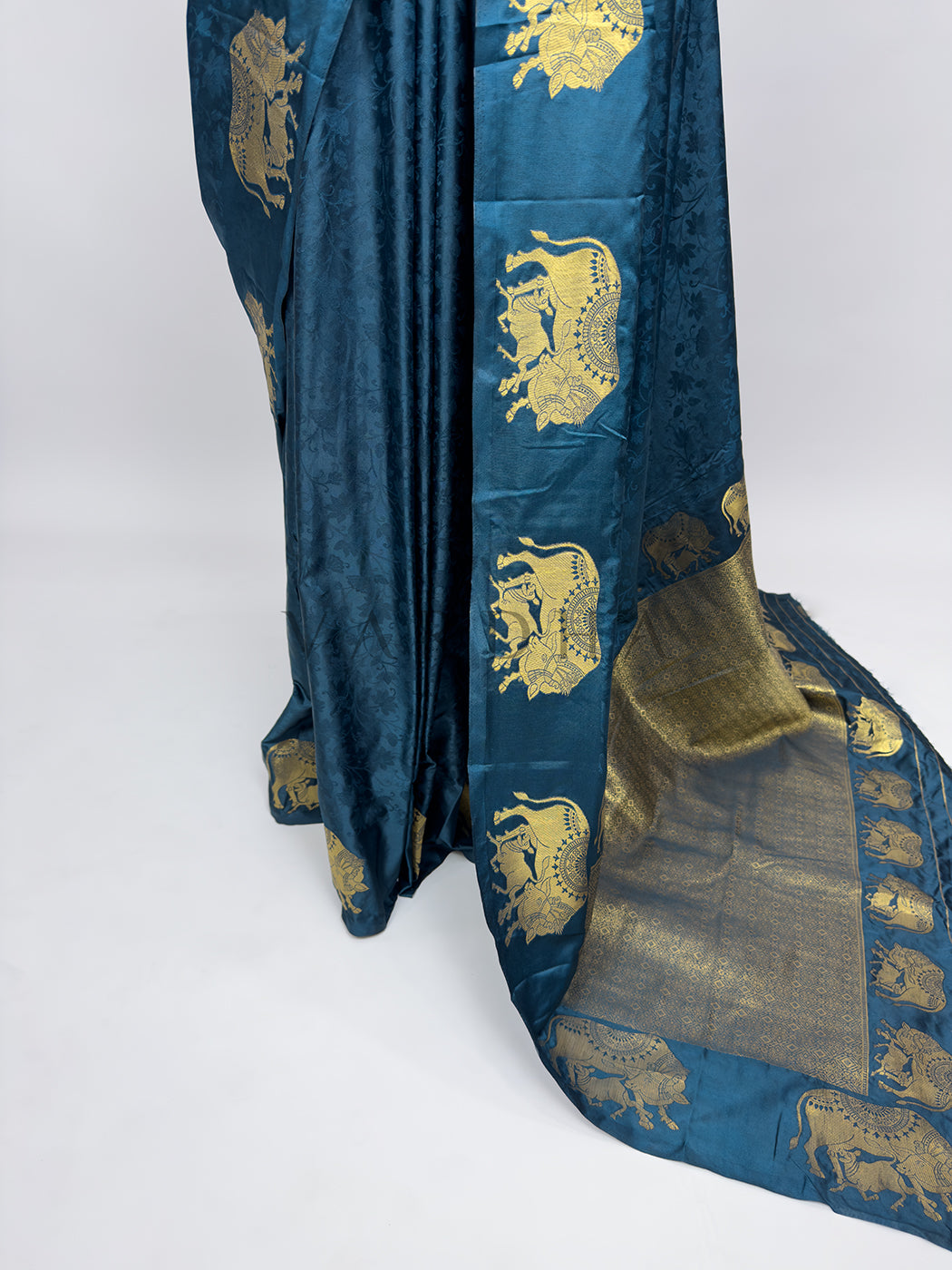 Blue fabric with gold embroidery on a white background A targeted view focusing on the lower drape and front pleats of the Midnight Blue Satin Silk Saree. The rich satin fabric creates smooth, lustrous vertical folds that catch the light, while a wide vertical panel decorated with a repeating line of detailed gold zari elephant motifs frames the side, flowing seamlessly into the grand pallu spread across the floor.