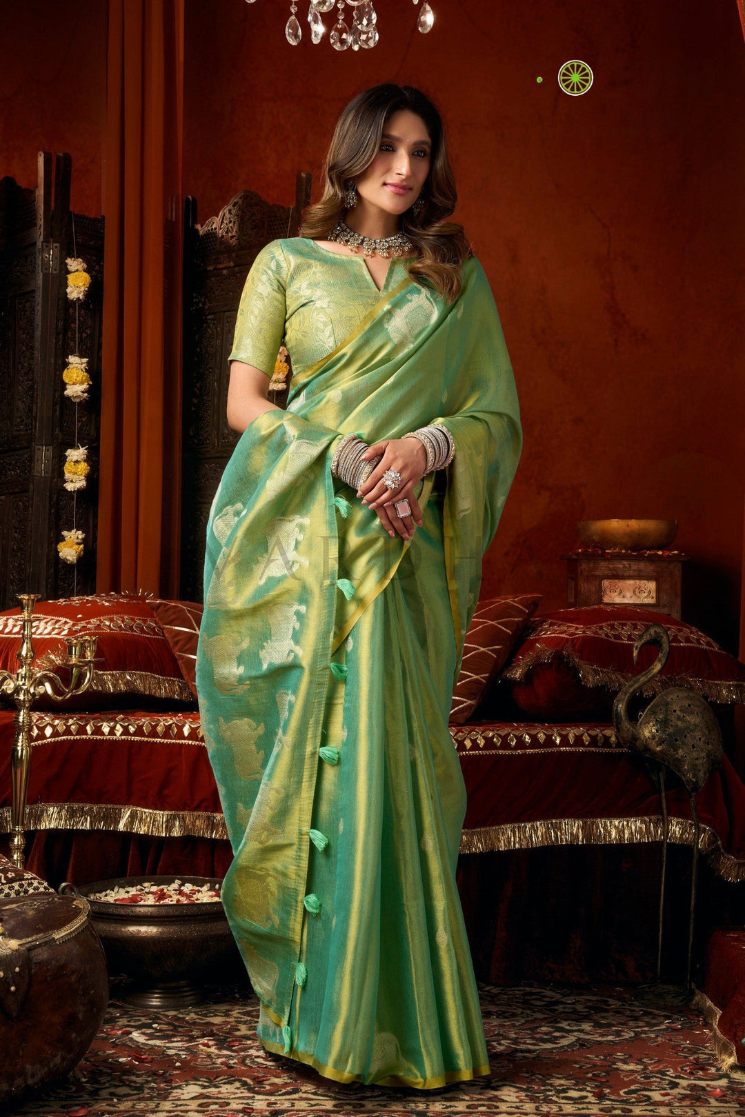 A woman wearing a light green silk saree with gold zari stripes and a matching green blouse, standing in an ornate traditional room with red drapery and warm brown walls. Stylish front drape of the pastel green heavy tissue silk saree showcasing its luxurious texture, perfect for mehendi and wedding wear.