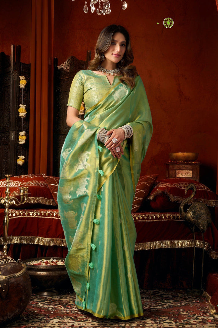 A woman wearing a light green silk saree with gold zari stripes and a matching green blouse, standing in an ornate traditional room with red drapery and warm brown walls. Stylish front drape of the pastel green heavy tissue silk saree showcasing its luxurious texture, perfect for mehendi and wedding wear.