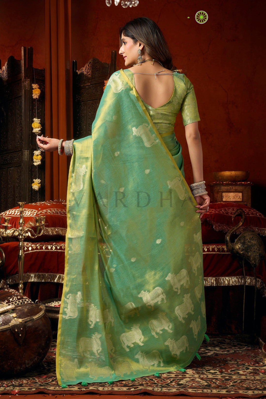 Woman in a green saree with elephant patterns in an indoor setting Back view of the radiant mint green tissue saree, emphasizing the rich golden metallic sheen, woven silver cow designs, and elegant fancy tassels.